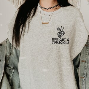 Puede incluir: Una sudadera gris con un gráfico de mano de esqueleto negro y el texto "Upright & Conscious".
