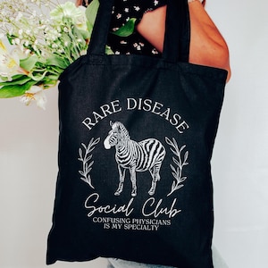 Może przedstawiać: Czarna torba typu tote z białym tekstem, który brzmi "Rare Disease Social Club Confusing Physicians is my specialty". W środku torby znajduje się biała ilustracja zebry.