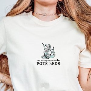 Puede incluir: Una camiseta blanca con una ilustración de cebra en blanco y negro en una pose de yoga. El texto "not everyone can be POTS HEDS" está impreso debajo de la cebra.