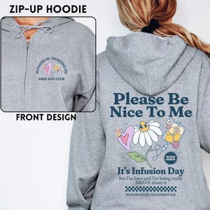 Może przedstawiać: Szara bluza z kapturem zapinana na zamek z przodu i z tyłu. Przedni wzór zawiera tekst i grafikę serca. Tylny wzór zawiera tekst "Please Be Nice To Me" i grafikę stokrotki. Bluza z kapturem zawiera również tekst "It's Infusion Day".