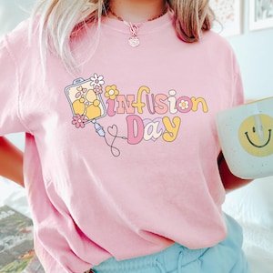 May include: A pink t-shirt featuring a graphic of a medical IV bag and the text "Infusion Day" in a retro font. The shirt is being worn by a person holding a smiley face mug.