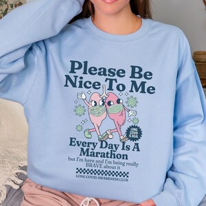 Puede incluir: Sudadera azul claro con un gráfico de dos pulmones con mascarillas y el texto "Please Be Nice To Me. Every Day Is A Marathon but I'm here and I'm being really BRAVE about it. LONG COVID AWARENESS CLUB."