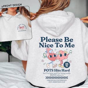 Pode incluir: Moletom cinza claro com a frase "Please Be Nice To Me" e um desenho animado de coração e cérebro. O design frontal apresenta o texto "Salty, Dizzy, Determined, POTS Warrior". O moletom também tem o texto "POTS Hits Hard".