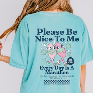 May include: A turquoise blue t-shirt with a graphic of two cartoon lungs wearing face masks and the text "Please Be Nice To Me". The graphic also includes the text "Every Day Is A Marathon but I'm here and I'm being really BRAVE about it" and "LONG COVID AWARENESS CLUB".