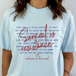 May include: Light blue t-shirt with red text that reads "they tell us to be patient. to wait for change to prove we are "disabled enough" to deserve care. to fight the same battles over and over. survival is just to exist. but every day we wake up. every time we refuse to be pushed aside. we are resisting. our survival is proof of our power. and we are not done fighting. ♡ we deserve more than survival"