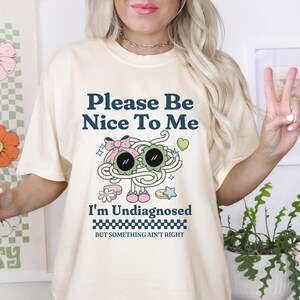 Puede incluir: Camiseta color crema con el texto "Please Be Nice To Me" sobre un dibujo animado de gafas de sol y espaguetis. Debajo del gráfico, el texto dice "I'm Undiagnosed" y "BUT SOMETHING AIN'T RIGHT".