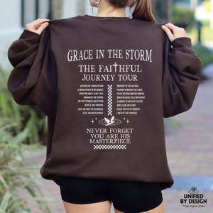 以下が含まれることがあります： 白文字で「Grace in the Storm, The Faithful Journey Tour」とインスピレーションを与えるフレーズのリストが書かれた茶色のスウェットシャツ。 「Never Forget You Are His Masterpiece」という文字も白で印刷されています。