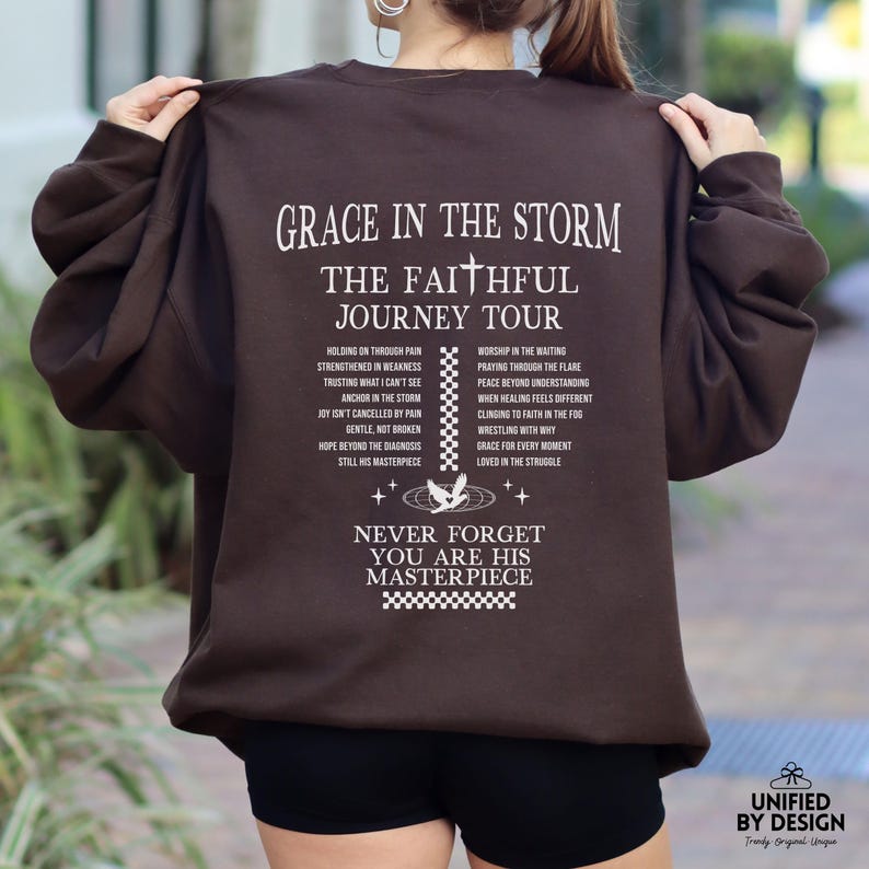 以下が含まれることがあります： 白文字で「Grace in the Storm, The Faithful Journey Tour」とインスピレーションを与えるフレーズのリストが書かれた茶色のスウェットシャツ。 「Never Forget You Are His Masterpiece」という文字も白で印刷されています。
