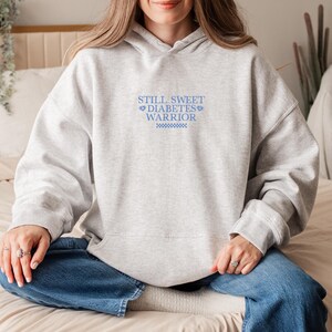 K&ouml;nnte beinhalten: Ein grauer Hoodie mit dem Text "STILL SWEET DIABETES WARRIOR" in blauer Schrift. Der Text ist mittig auf dem Hoodie platziert und von zwei kleinen Herzen umgeben.