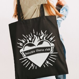 May include: Black tote bag with a white graphic of a heart on fire with the text "invisible illness club" on a banner.