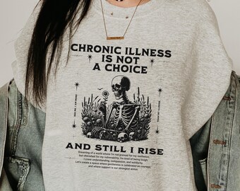 Puede incluir: Sudadera gris con un gráfico en blanco y negro de un esqueleto sosteniendo una flor. El texto "Chronic Illness is not a choice and still I rise" está impreso en la camiseta.