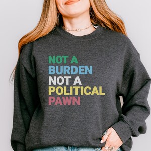 May include: A dark grey sweatshirt with the text "NOT A BURDEN NOT A POLITICAL PAWN" in colorful letters.