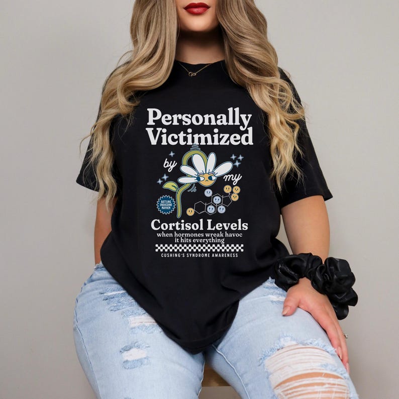 May include: Black t-shirt with white text that reads "Personally Victimized by my Cortisol Levels" with a cartoon daisy and the text "Cushing's Syndrome Awareness".