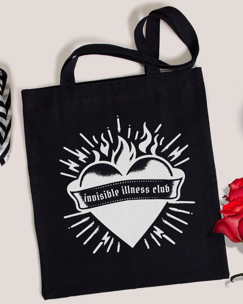 Black tote bag with a tattoo inspired flaming heart with ribbon banner across the middle that says, "invisible illness club"