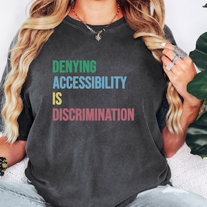 May include: A dark gray t-shirt with the text "DENYING ACCESSIBILITY IS DISCRIMINATION" in colorful letters.
