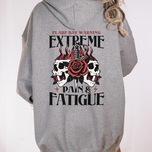 Puede incluir: Sudadera gris con un estampado gráfico de dos calaveras con una rosa y una daga entre ellas. El texto "Flare Day Warning Extreme Pain & Fatigue" está impreso debajo del gráfico.