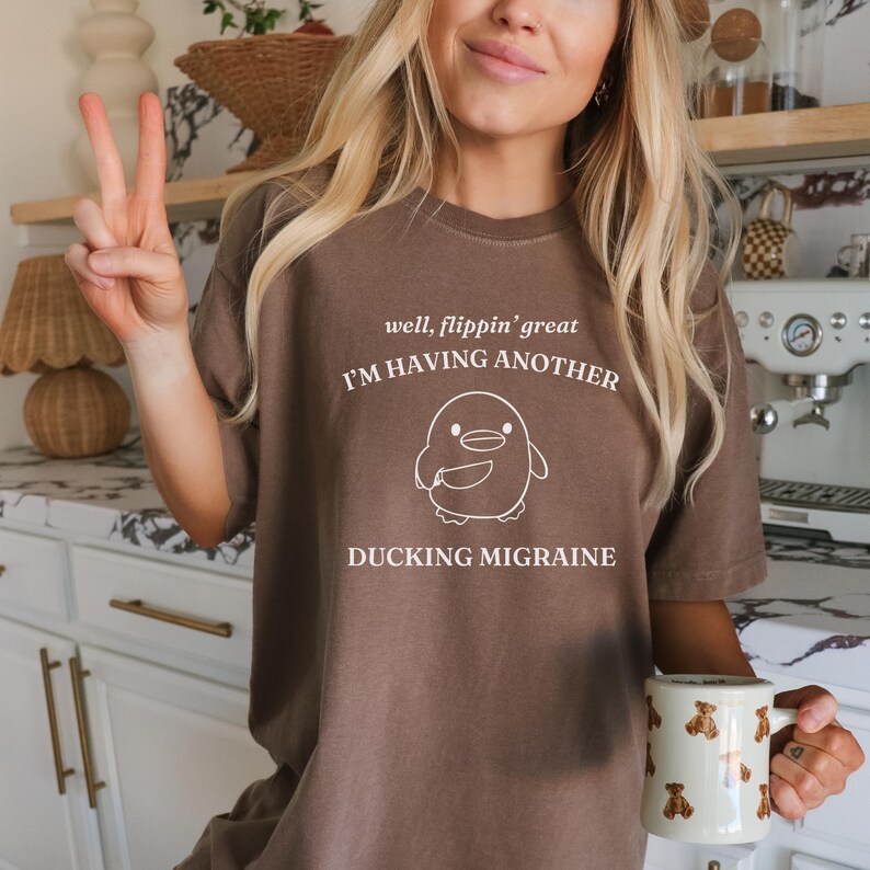 May include: A brown t-shirt with a white graphic of a penguin holding a cup and the text "well, flippin' great I'M HAVING ANOTHER DUCKING MIGRAINE".