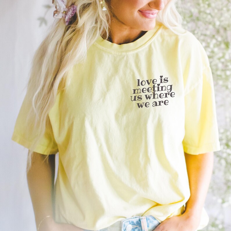 Op de afbeelding: Een lichtgeel t-shirt met de tekst "love is meeting us where we are" in het zwart gedrukt. Het shirt is gemaakt van een zacht, comfortabel materiaal.