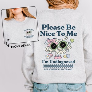 Puede incluir: Una sudadera gris claro con el texto "Please Be Nice To Me" e "I'm Undiagnosed" en azul marino. La parte trasera presenta un diseño de dibujos animados de espaguetis con gafas de sol y un lazo. La parte delantera tiene una versión más pequeña del diseño.