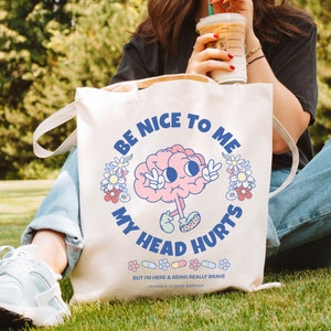 Puede incluir: Bolsa de tela blanca con un cerebro de dibujos animados que lleva un signo de paz y el texto "Be Nice To Me My Head Hurts" con una fila de p&iacute;ldoras y el texto "But I'm here & being really brave Invisible Illness Warrior".