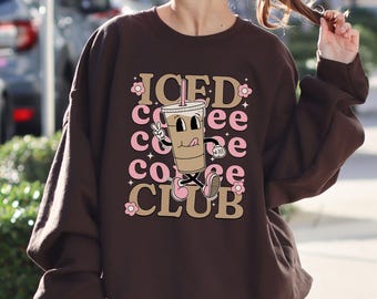 Op de afbeelding: Een bruine sweatshirt met de tekst "ICED COFFEE CLUB" in een retro lettertype. Het ontwerp bevat een cartoon koffiekopje met een vredesteken en roze accenten.