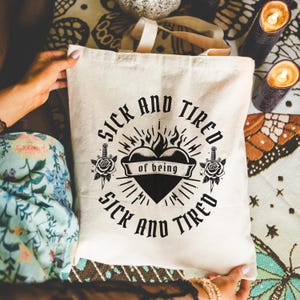 Könnte beinhalten: Eine weiße Canvas-Tragetasche mit einem schwarzen grafischen Design. Die Grafik zeigt ein Herz mit Flammen, zwei Dolche und den Text "Sick and Tired of being Sick and Tired".