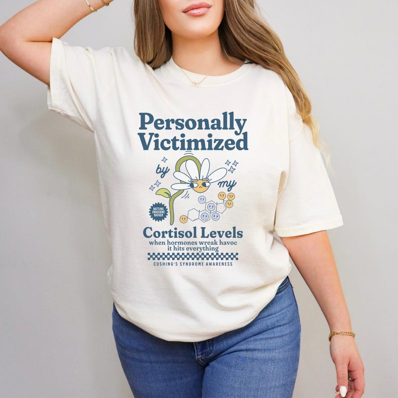 May include: A white t-shirt with a graphic design that says "Personally Victimized by my Cortisol Levels" with a cartoon daisy and a chemical structure. The text below the graphic says "when hormones wreak havoc it hits everything" and "Cushing's Syndrome Awareness".