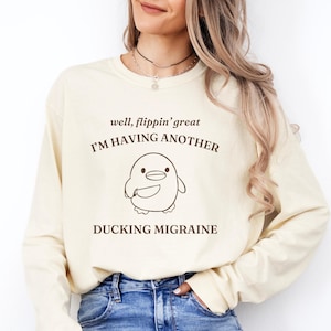 Puede incluir: Una camiseta de manga larga de color crema con un gráfico marrón de un pingüino sosteniendo un cuchillo. El texto "well, flippin' great I'M HAVING ANOTHER DUCKING MIGRAINE" está impreso en la camiseta.