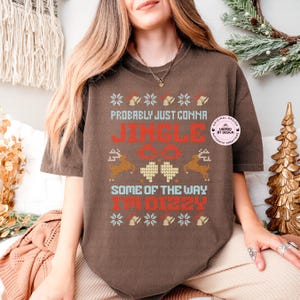 Peut inclure: Un t-shirt marron avec un motif festif. Le t-shirt pr&eacute;sente le texte "Probably Just Comma Jingle All The Way I'm Dizzy" en rouge et blanc, ainsi que des graphiques de rennes et de flocons de neige. Le t-shirt a une coupe d&eacute;contract&eacute;e.