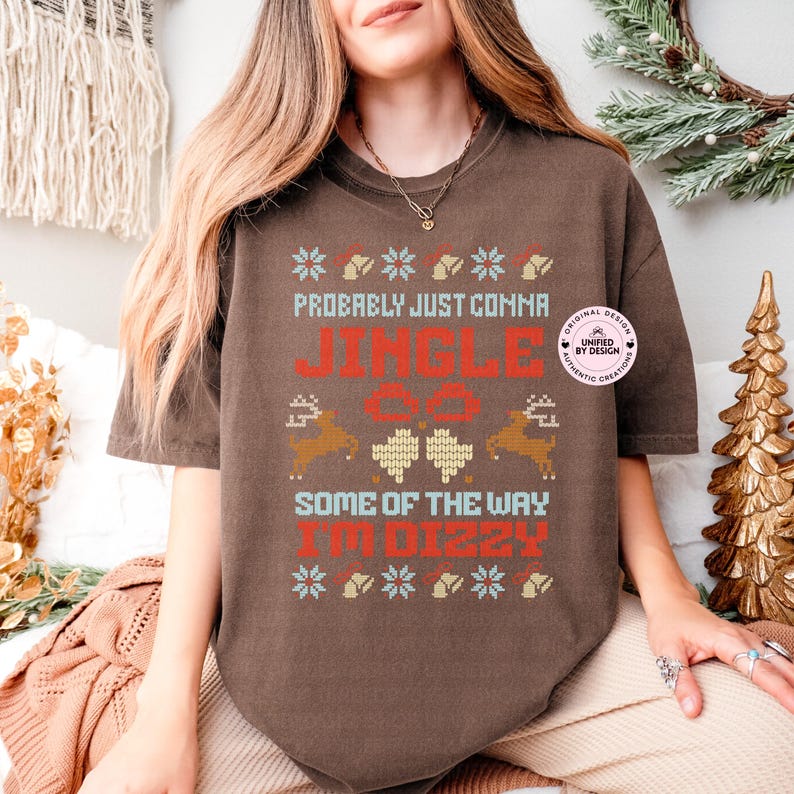 Peut inclure: Un t-shirt marron avec un motif festif. Le t-shirt pr&eacute;sente le texte "Probably Just Comma Jingle All The Way I'm Dizzy" en rouge et blanc, ainsi que des graphiques de rennes et de flocons de neige. Le t-shirt a une coupe d&eacute;contract&eacute;e.