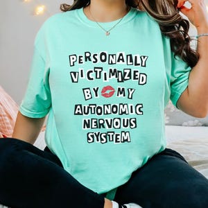 May include: A turquoise t-shirt with the text "PERSONALLY VICTIMIZED BY 💋 MY AUTONOMIC NERVOUS SYSTEM" printed in black.