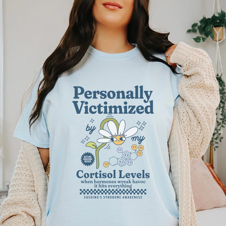 May include: Light blue t-shirt with a graphic design of a daisy with a face and the text "Personally Victimized by my Cortisol Levels when hormones wreak havoc it hits everything Cushing's Syndrome Awareness".