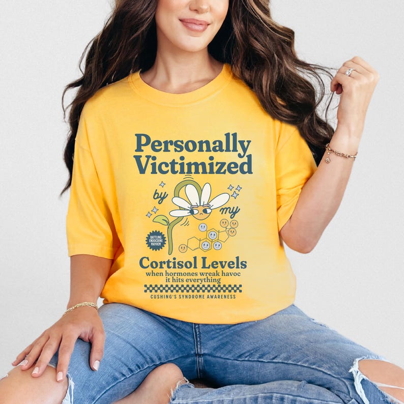 May include: Yellow t-shirt with a graphic of a daisy with a face and the text "Personally Victimized by my Cortisol Levels" and "Cushing's Syndrome Awareness".