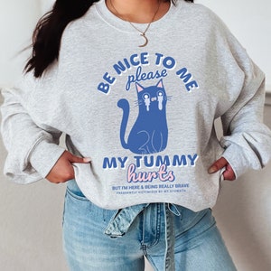 Op de afbeelding: Grijs sweatshirt met een blauwe kat illustratie en de tekst "BE NICE TO ME please MY TUMMY hurts BUT I'M HERE & BEING REALLY BRAVE FREQUENTLY VICTIMIZED BY MY STOMACH".