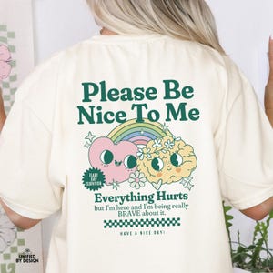 May include: A cream colored t-shirt with a green graphic that says "Please Be Nice To Me" with a rainbow, a heart, and a brain with flowers. The graphic also says "Everything Hurts but I'm here and I'm being really BRAVE about it. Have a nice day!"