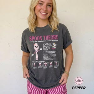 May include: Dark gray t-shirt with the text "SPOON THEORY" in pink. The shirt features a cartoon spoon character and the phrase "It's More Than Just Feeling Tired". The shirt also has a graphic of daisies.