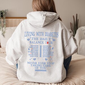 K&ouml;nnte beinhalten: Grauer Hoodie mit einem blauen und roten Grafikdruck, der "Leben mit Diabetes + Die t&auml;gliche Balance-Tour" sagt und t&auml;gliche Aufgaben im Zusammenhang mit der Diabetes-Behandlung auflistet. Die Grafik enth&auml;lt auch den Text "H&ouml;r niemals auf, auf dich aufzupassen".