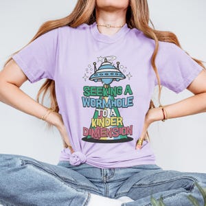 May include: A purple t-shirt with a graphic of a UFO and the text "Seeking a Wormhole to a Kinder Dimension".