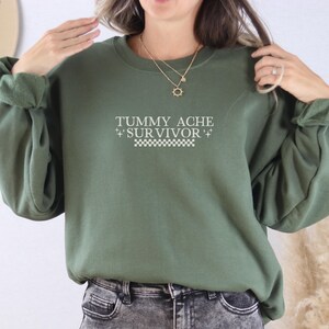 May include: An olive green sweatshirt with the text "TUMMY ACHE + SURVIVOR +" in white. The text is centered on the front of the sweatshirt, above a white and black checkered pattern. The sweatshirt has long sleeves.