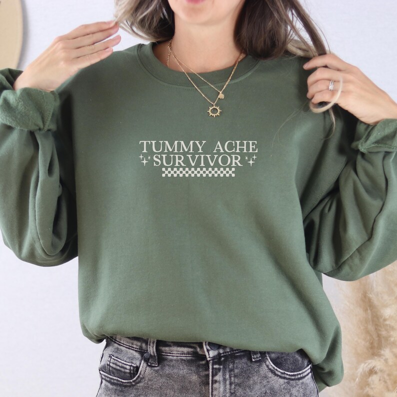 May include: An olive green sweatshirt with the text "TUMMY ACHE + SURVIVOR +" in white. The text is centered on the front of the sweatshirt, above a white and black checkered pattern. The sweatshirt has long sleeves.