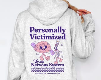 Op de afbeelding: Grijze hoodie met een paarse afbeelding van een brein en een ruggengraat die vechten met bokshandschoenen. De tekst luidt "Persoonlijk slachtoffer van mijn zenuwstelsel en het verwart knuffels met stoten" en "Fibromyalgia Awareness Club".