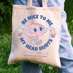 Puede incluir: Una bolsa de tela beige con un cerebro de dibujos animados que lleva un signo de paz y el texto "S&eacute; amable conmigo, me duele la cabeza". La bolsa tambi&eacute;n tiene una fila de p&iacute;ldoras coloridas y flores con el texto "Pero estoy aqu&iacute; y siendo realmente valiente Guerrero de la enfermedad invisible".