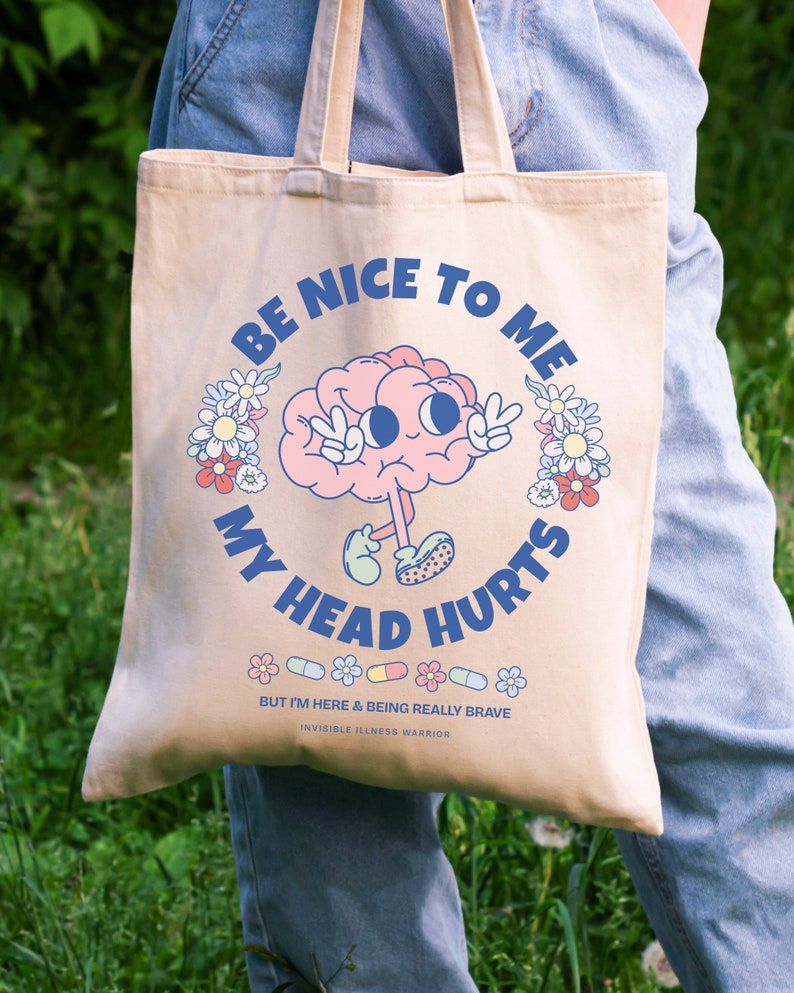 Puede incluir: Una bolsa de tela beige con un cerebro de dibujos animados que lleva un signo de paz y el texto "S&eacute; amable conmigo, me duele la cabeza". La bolsa tambi&eacute;n tiene una fila de p&iacute;ldoras coloridas y flores con el texto "Pero estoy aqu&iacute; y siendo realmente valiente Guerrero de la enfermedad invisible".
