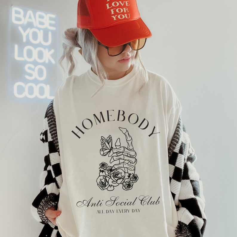 Tshirt with a drawing of a skeleton hand accented by flowers and a butterfly. Text says Homebody Anti Social Club - All Day Every Day