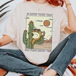 Op de afbeelding: Een beige T-shirt met een cartoonillustratie van een kikker die een zonnebril draagt en ontspant op een cactus. De tekst "NO ROOTIN' TOOTIN' TODAY" staat boven de afbeelding en "ONLY TUMMY HURTIN' VIBES" eronder.