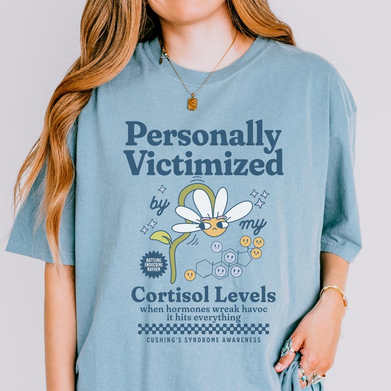 May include: A light blue t-shirt with a graphic design of a daisy with a sad face and the text "Personally Victimized by my Cortisol Levels". The text "Battling Endocrine Mayhem" and "Cushing's Syndrome Awareness" are also included in the design.