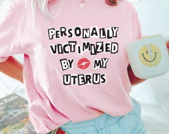 Puede incluir: Camiseta rosa con el texto "PERSONALLY VICTIMIZED BY MY UTERUS" en letras negras con una impresión de labios rojos entre las palabras "BY" y "MY".