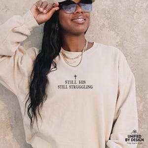以下が含まれることがあります： 十字架のシンボルと「Still His Still Struggling」というテキストが書かれたベージュのトレーナー。ゆったりとしたフィット感とクルーネックです。