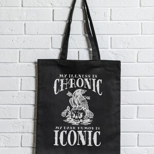 May include: Black tote bag with white text and graphic. The text reads "MY ILLNESS IS CHRONIC" above a crow perched on a skull with arrows, and "MY DARK HUMOR IS ICONIC" below. The bag has black handles.