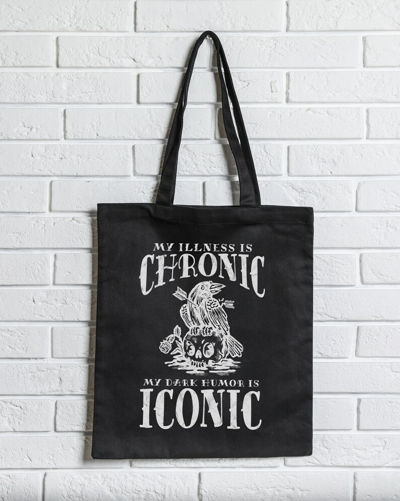 May include: Black tote bag with white text and graphic. The text reads "MY ILLNESS IS CHRONIC" above a crow perched on a skull with arrows, and "MY DARK HUMOR IS ICONIC" below. The bag has black handles.
